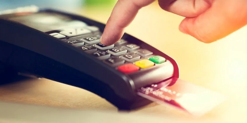 Dedo digitando a senha numa maquininha de cartão com um cartão inserido
