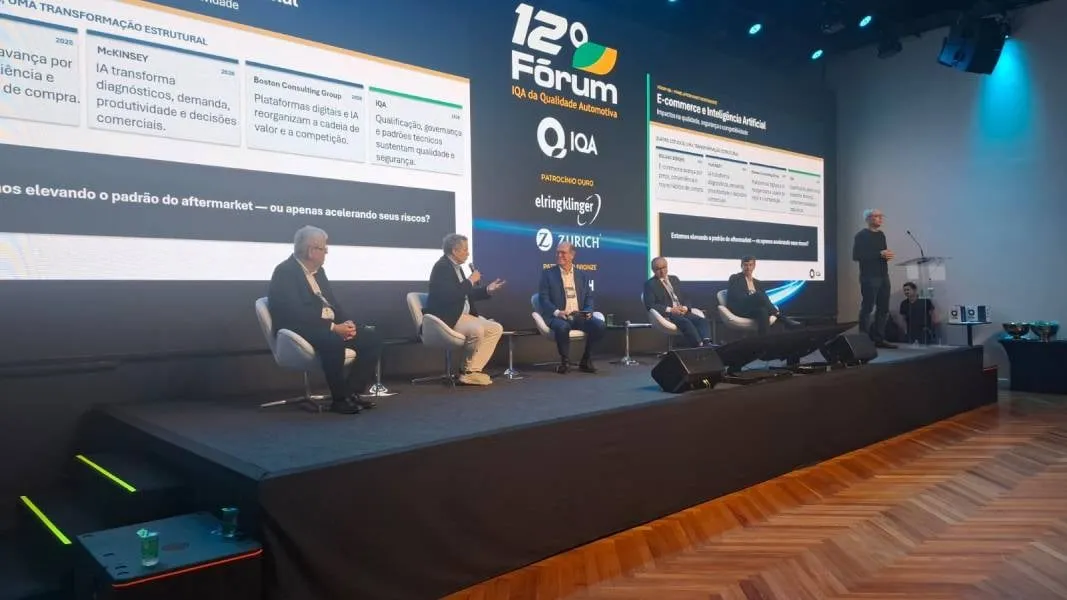 Painel de debate no palco do 12º Fórum IQA da Qualidade Automotiva, com cinco participantes sentados, um apresentador em pé e telões ao fundo