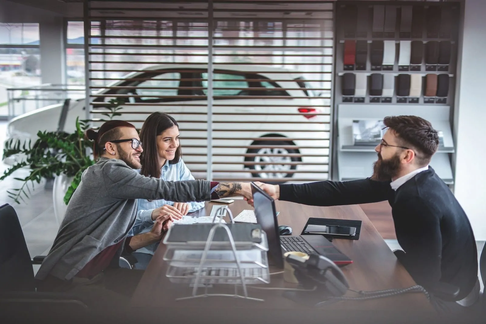 Vendedor apertando a mão de um casal de clientes sorridentes numa mesa de concessionária, com um carro branco ao fundo