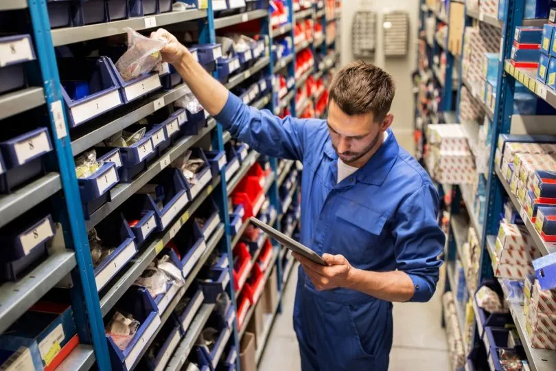Empleado con mameluco azul consultando una tablet entre estanterías de piezas en el depósito de una casa de repuestos