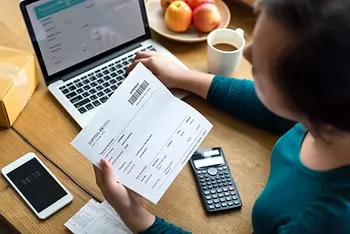 Pessoa segurando um boleto de pagamento diante de um notebook, com celular e calculadora sobre a mesa